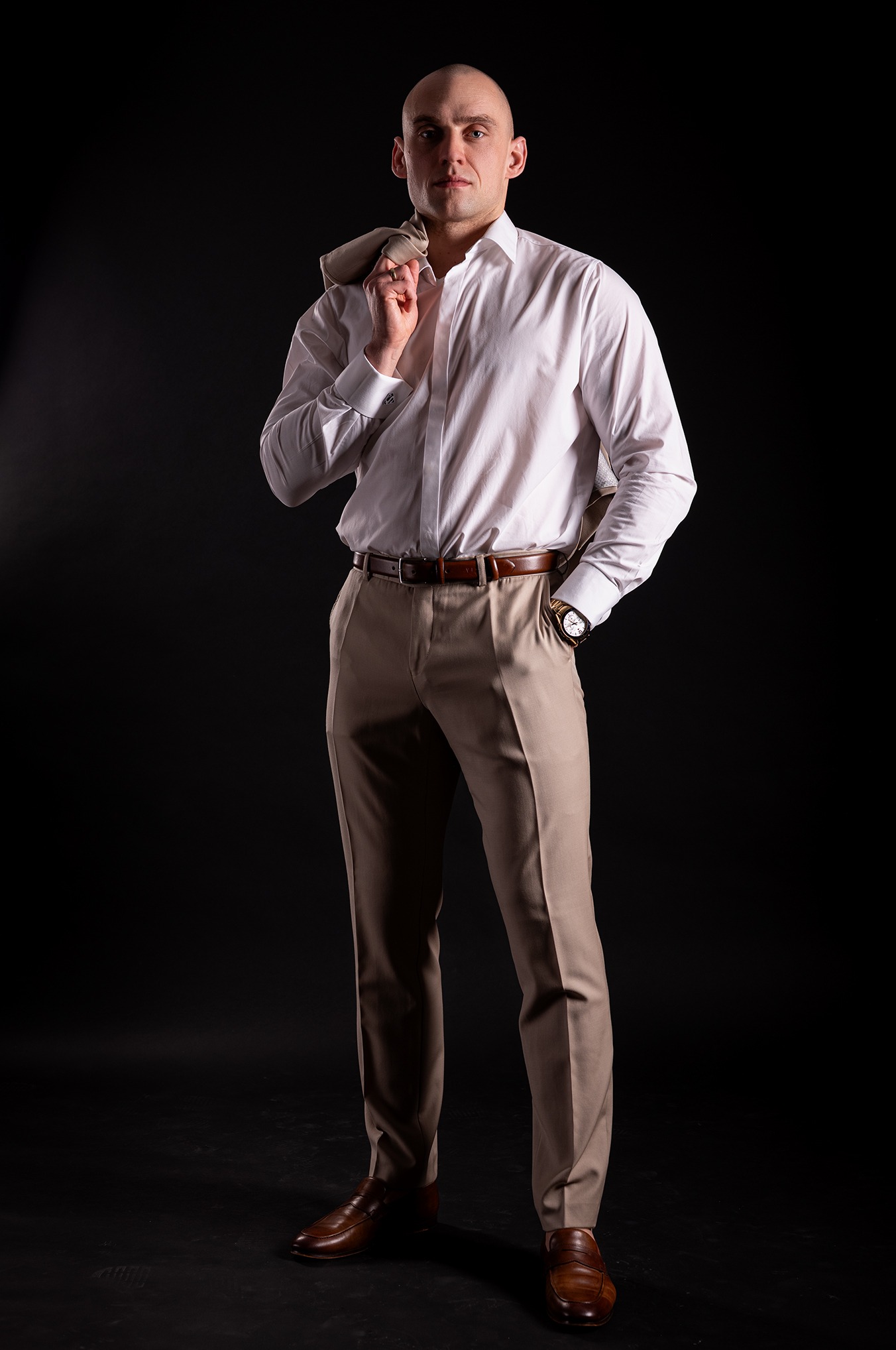 Mężczyzna w białej koszuli i beżowych spodniach, z marynarką na ramieniu, pozuje na ciemnym tle, prezentując elegancki styl i zegarek na nadgarstku.