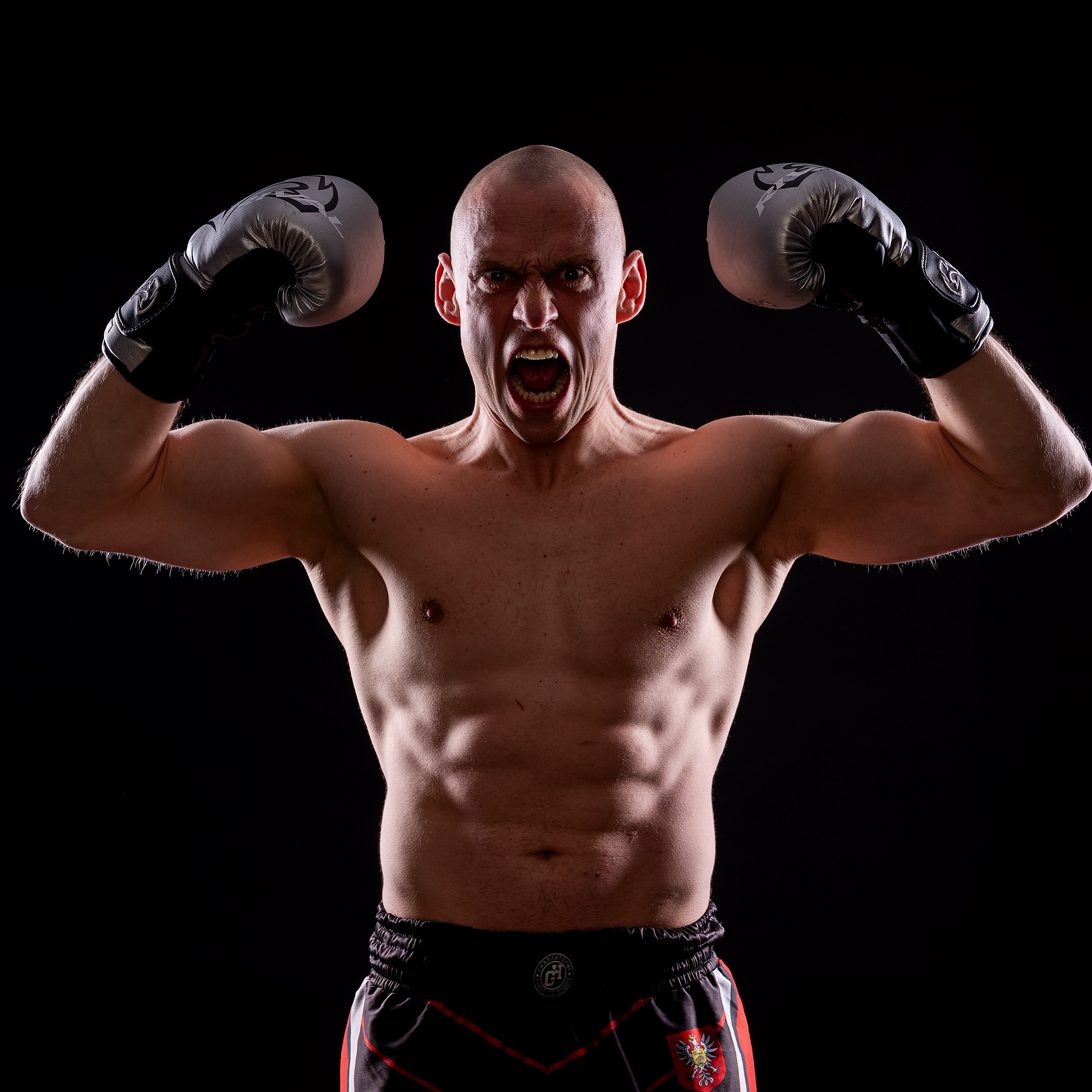 Intensywny portret mężczyzny w rękawicach bokserskich, z napiętymi mięśniami i krzykiem, na czarnym tle. Dynamiczne ujęcie podkreśla siłę i determinację.