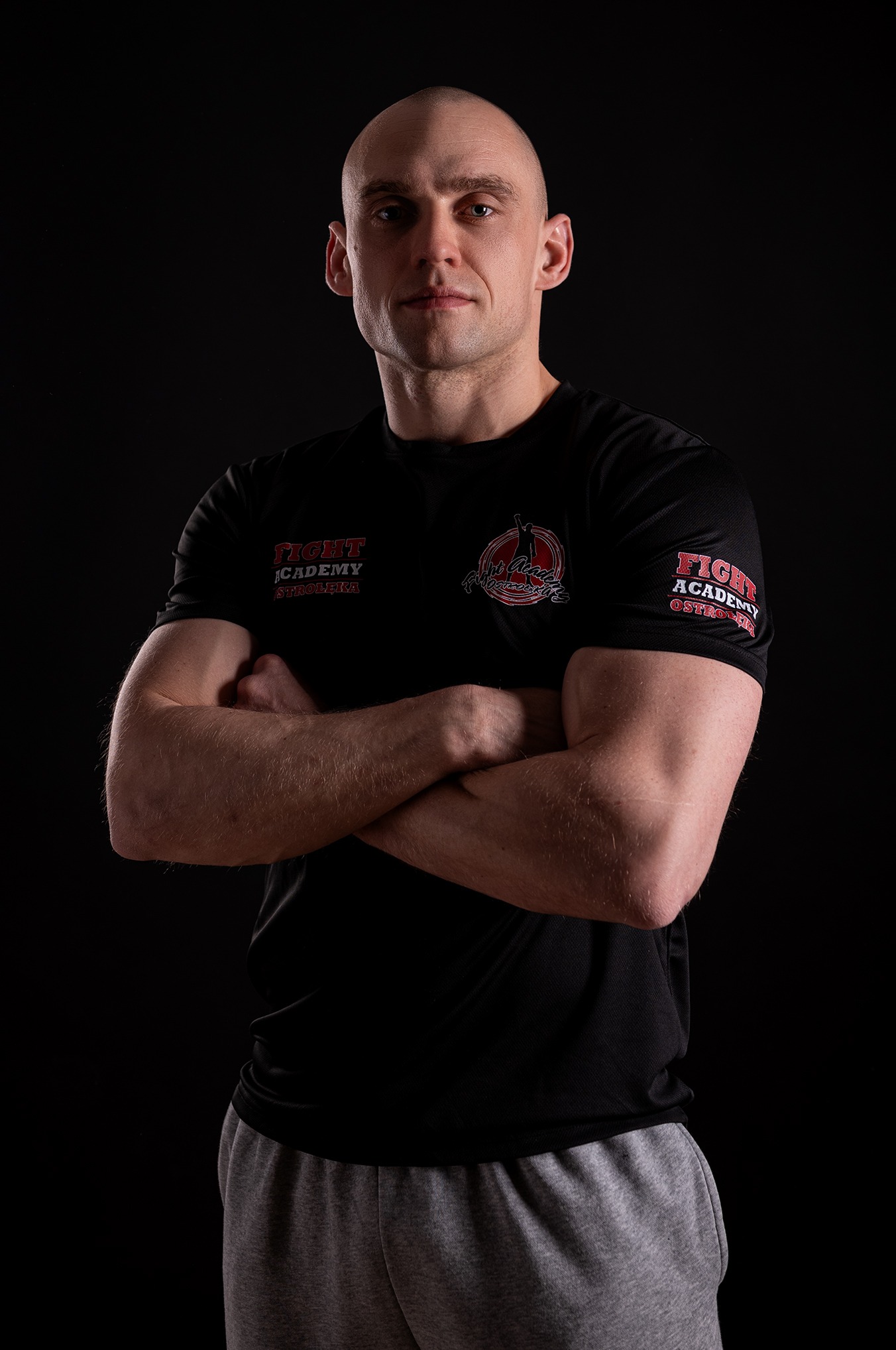 Portret mężczyzny na czarnym tle, z założonymi rękami, w czarnej koszulce z logo 'Fight Academy Ostrołęka', z widocznym oświetleniem studyjnym.
