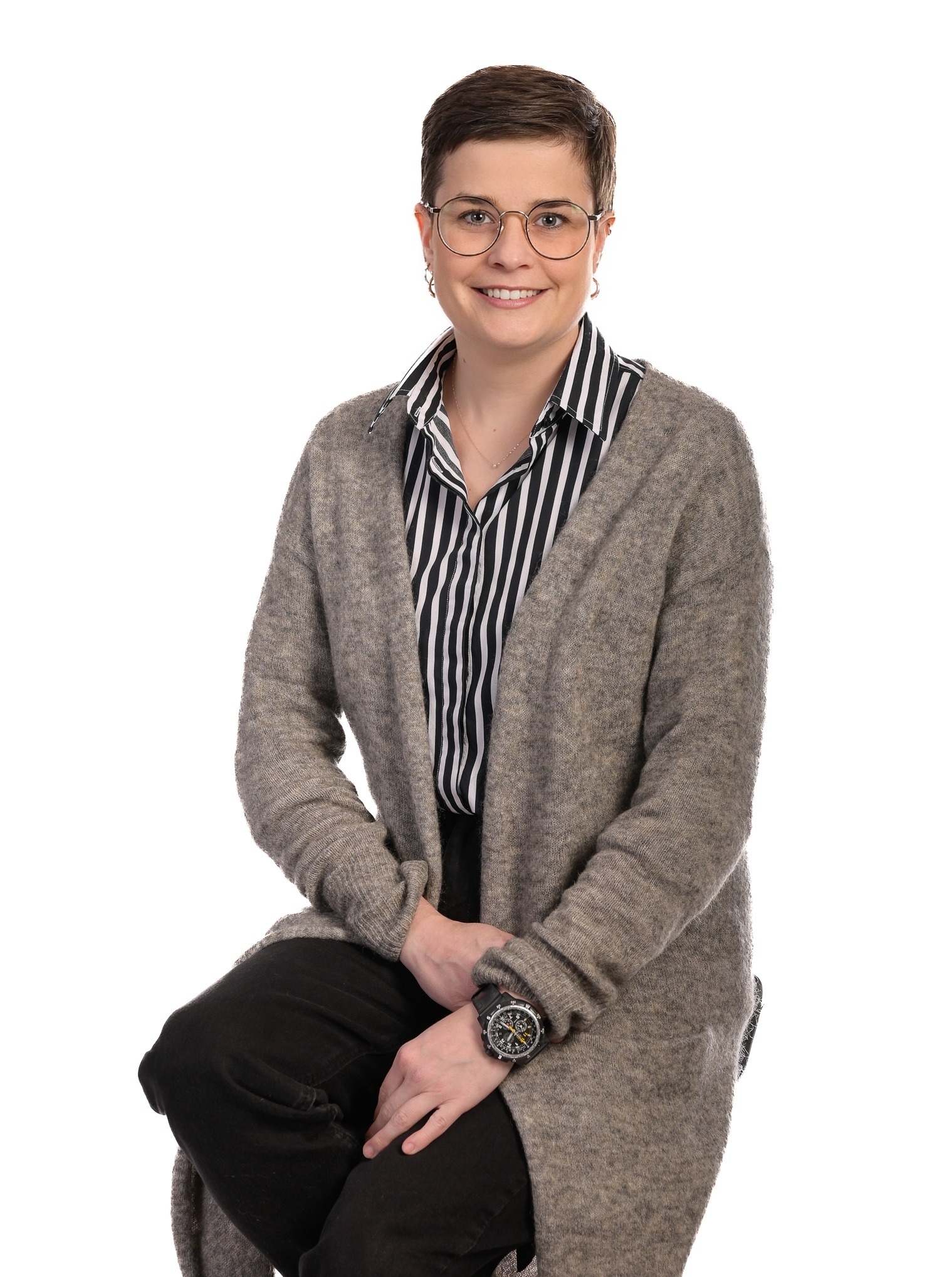 Portret uśmiechniętej kobiety w okularach, siedzącej na krześle, w swetrze i koszuli w paski, na białym tle. Widoczny zegarek na ręku.