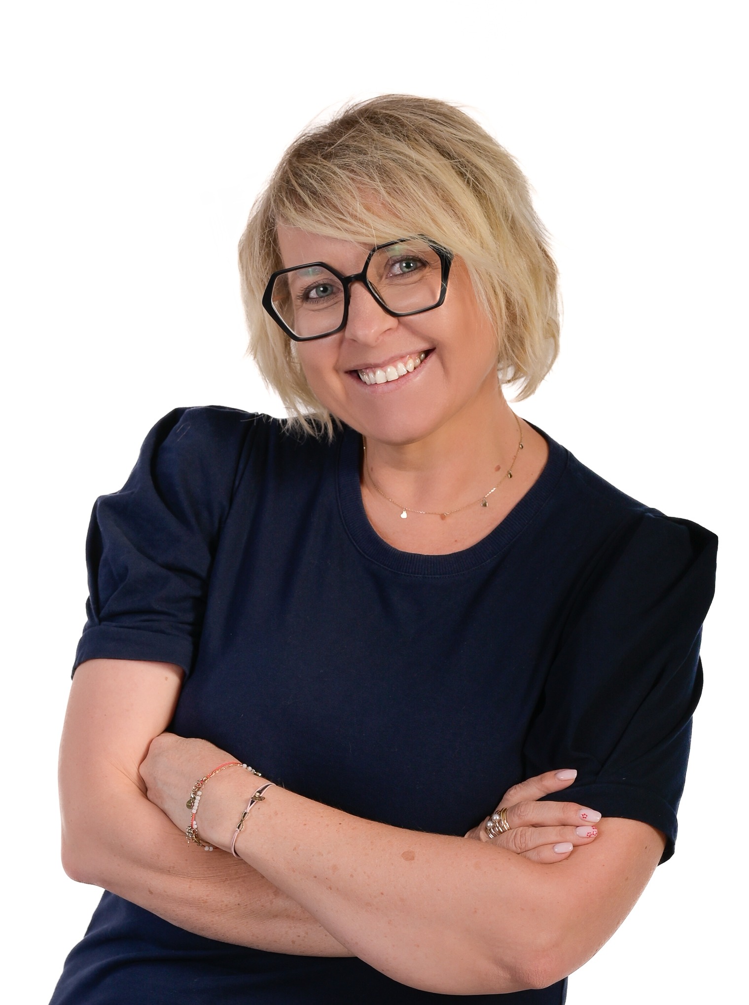 Uśmiechnięta blond kobieta w okularach i granatowej koszulce, z założonymi rękami na białym tle. Portret.