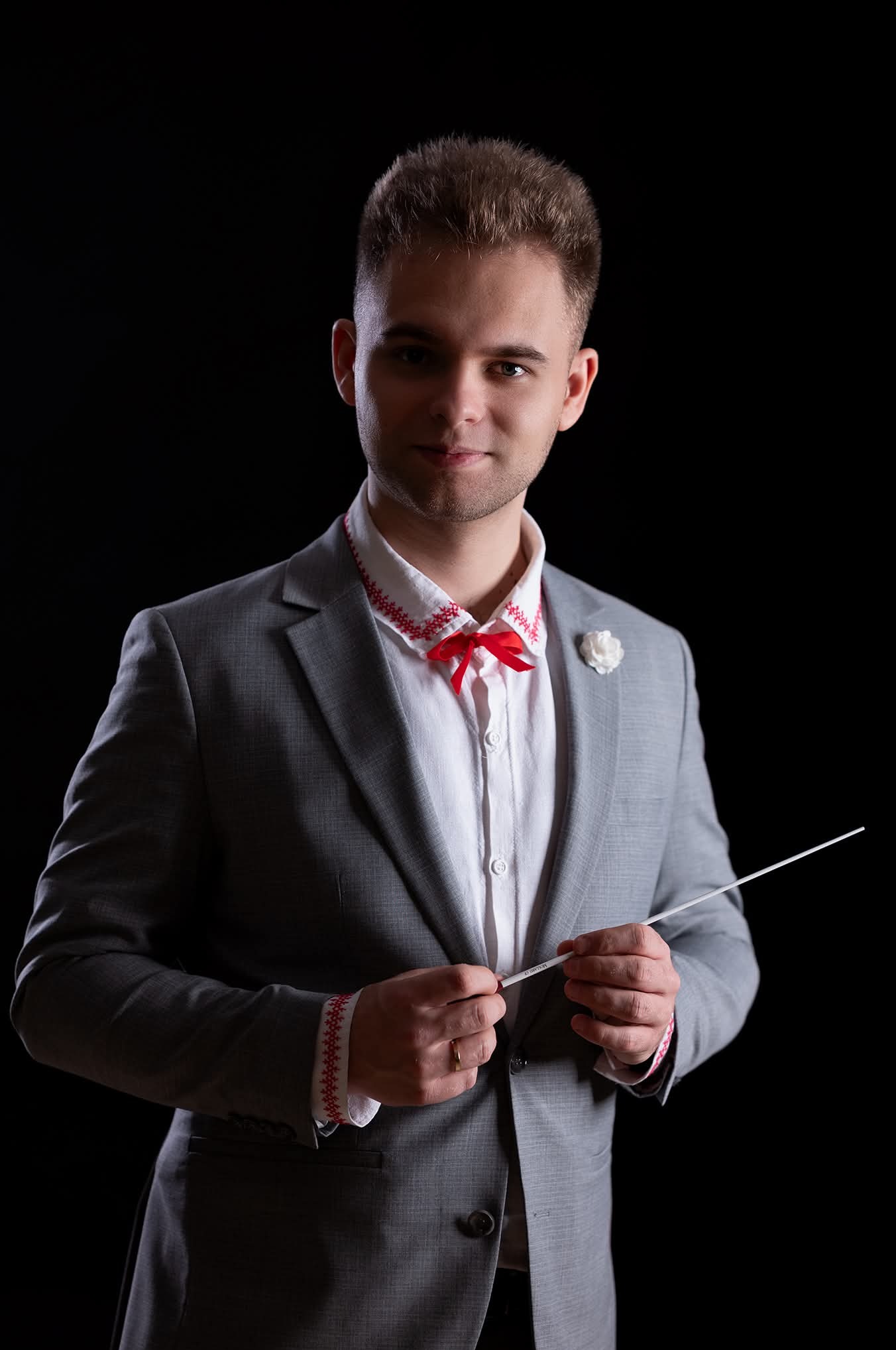 Portret mężczyzny w szarym garniturze z czerwoną muchą i haftowaną koszulą, trzymającego batutę na czarnym tle. Wyjątkowe oświetlenie podkreśla detale stroju.