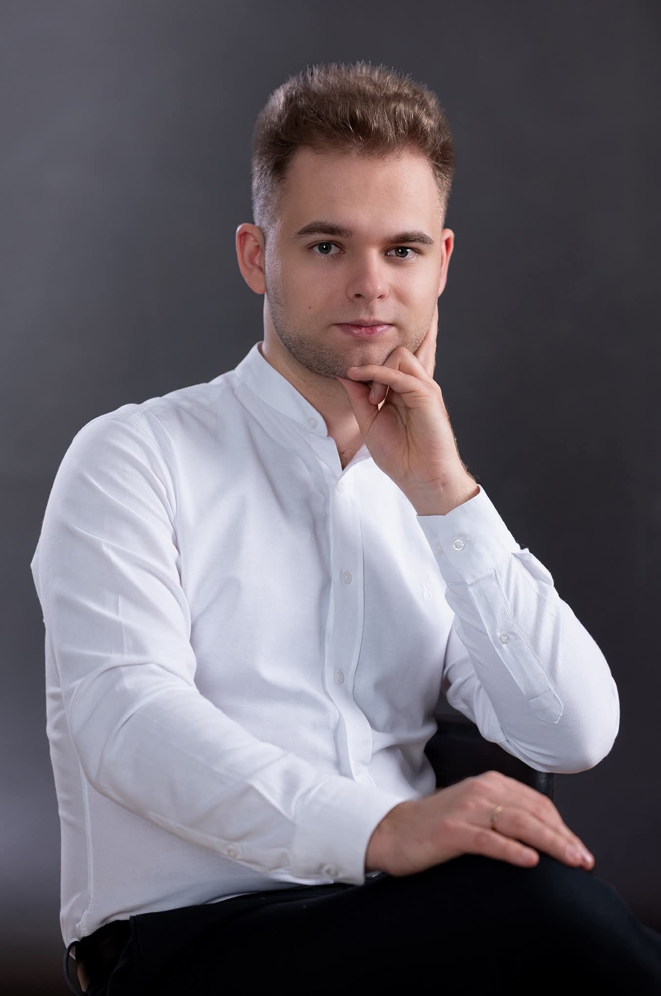 Mężczyzna w białej koszuli, z ręką pod brodą, pozuje na ciemnoszarym tle. Ujęcie portretowe, z delikatnym uśmiechem i pierścionkiem na palcu.