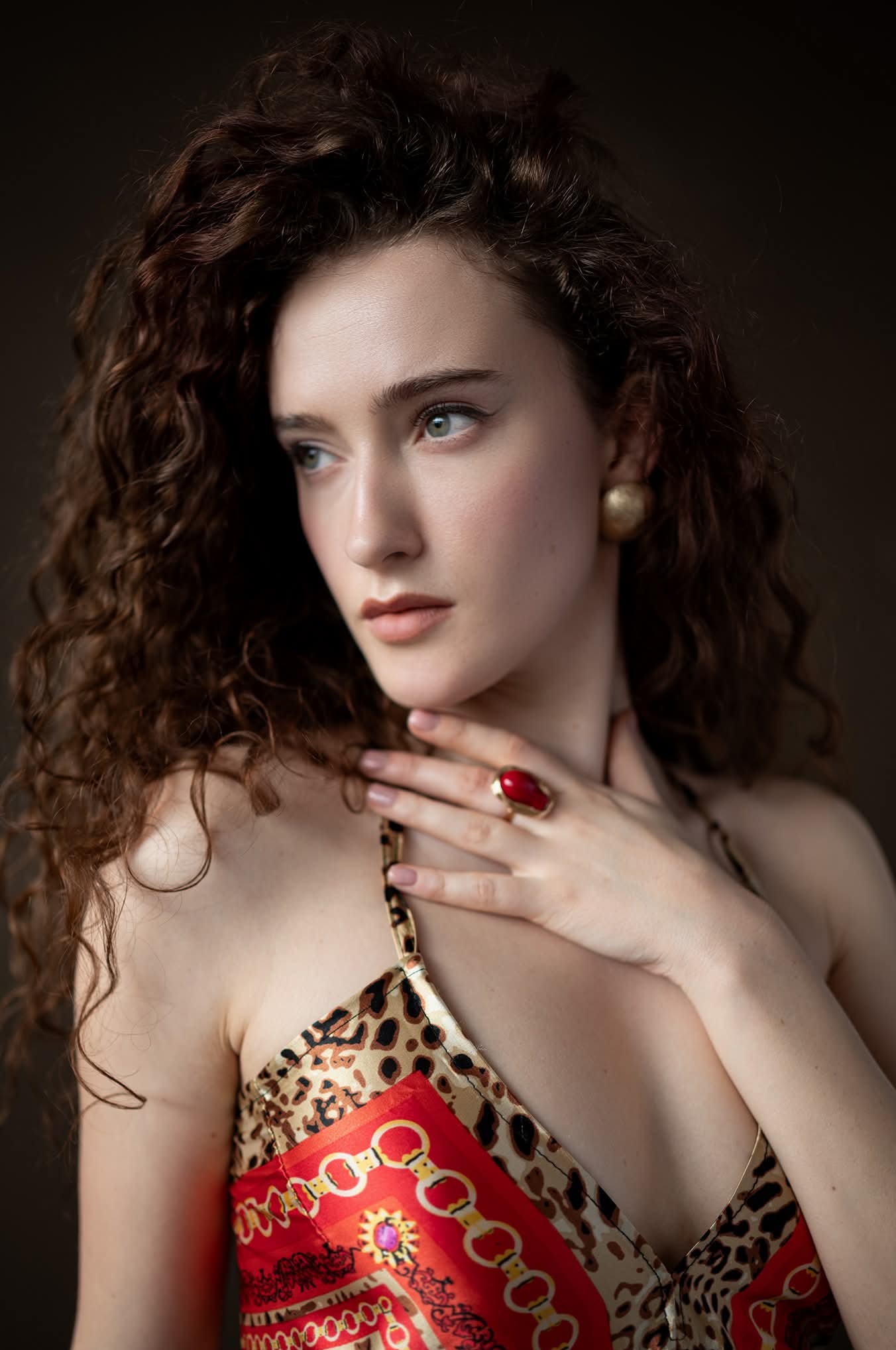 Portret kobiety o kręconych włosach, z delikatnym makijażem, w bluzce z wzorem panterki i czerwonym pierścionkiem, pozującej na ciemnym tle.