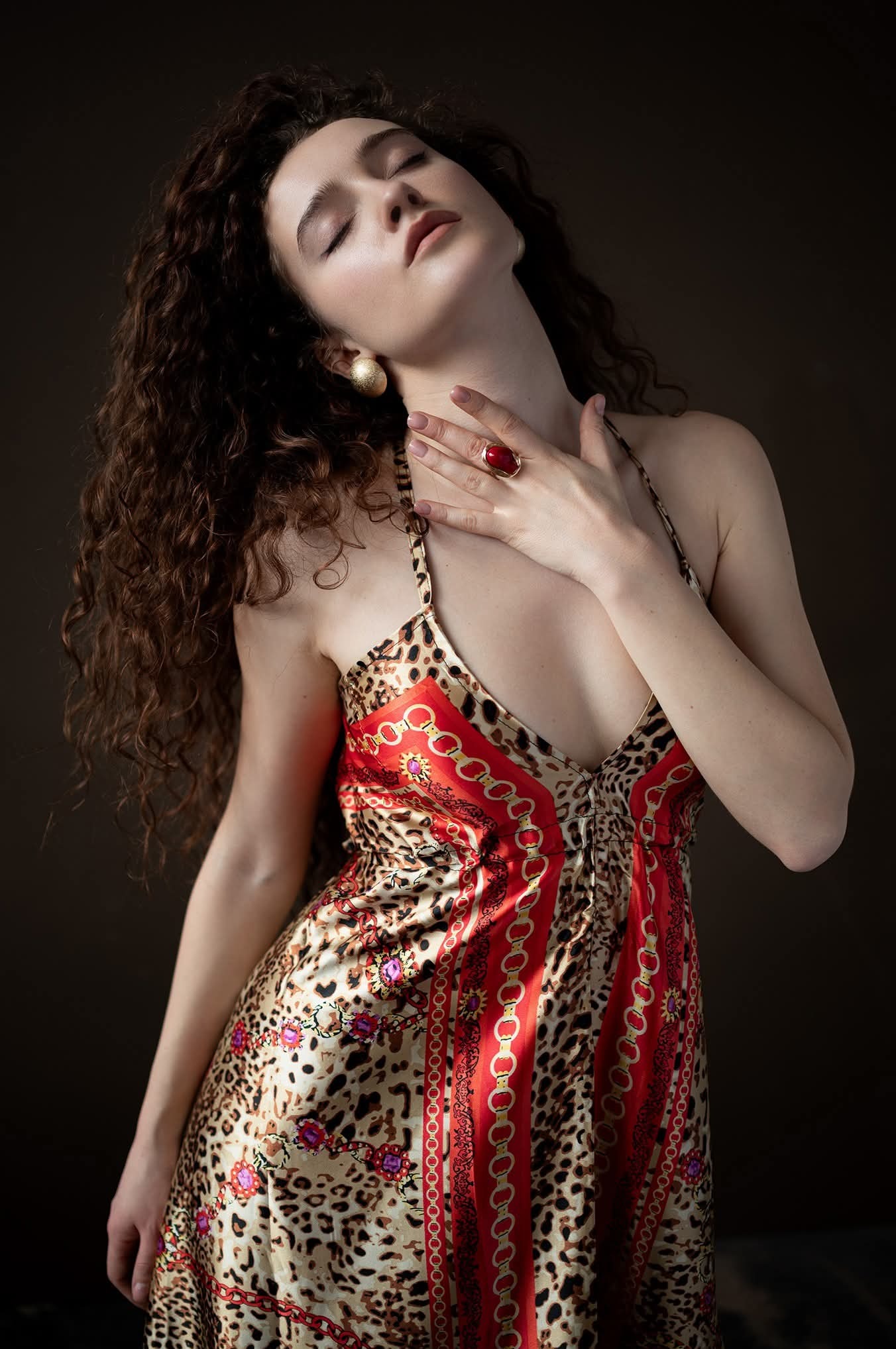 Portret kobiety z kręconymi włosami w sukience w panterkę, z zamkniętymi oczami, w złotych kolczykach i pierścionku z czerwonym kamieniem na ciemnym tle.