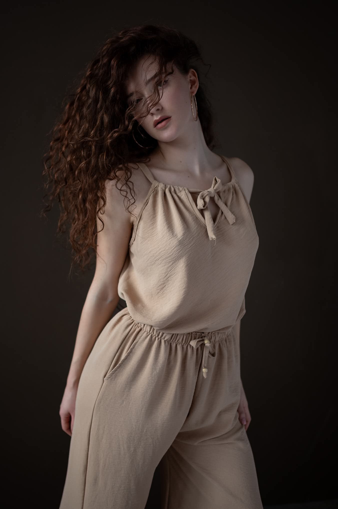 Portret kobiety z burzą loków, ubrana w beżowy kombinezon. Delikatne światło podkreśla naturalność, a ciemne tło dodaje głębi. Uwagę przyciągają złote kolczyki.