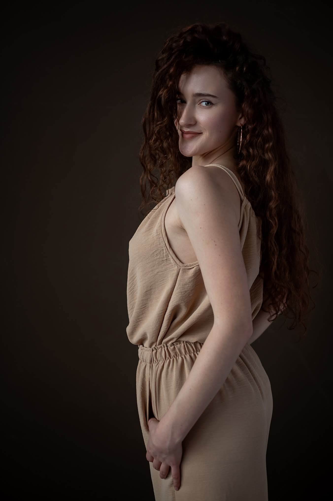 Portret kobiety z burzą rudych loków, ubrana w beżowy kombinezon, spogląda przez ramię z delikatnym uśmiechem na ciemnym, jednolitym tle. Złote kolczyki w kształcie długich prostokątów.