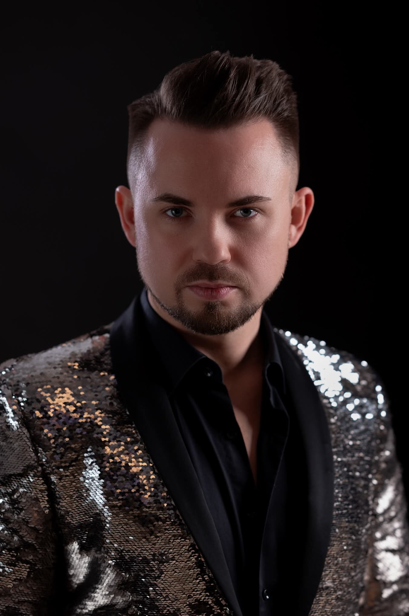 Portret mężczyzny z krótką fryzurą, w czarnej koszuli i cekinowej marynarce na czarnym tle, z poważnym wyrazem twarzy i intensywnym spojrzeniem w obiektyw.