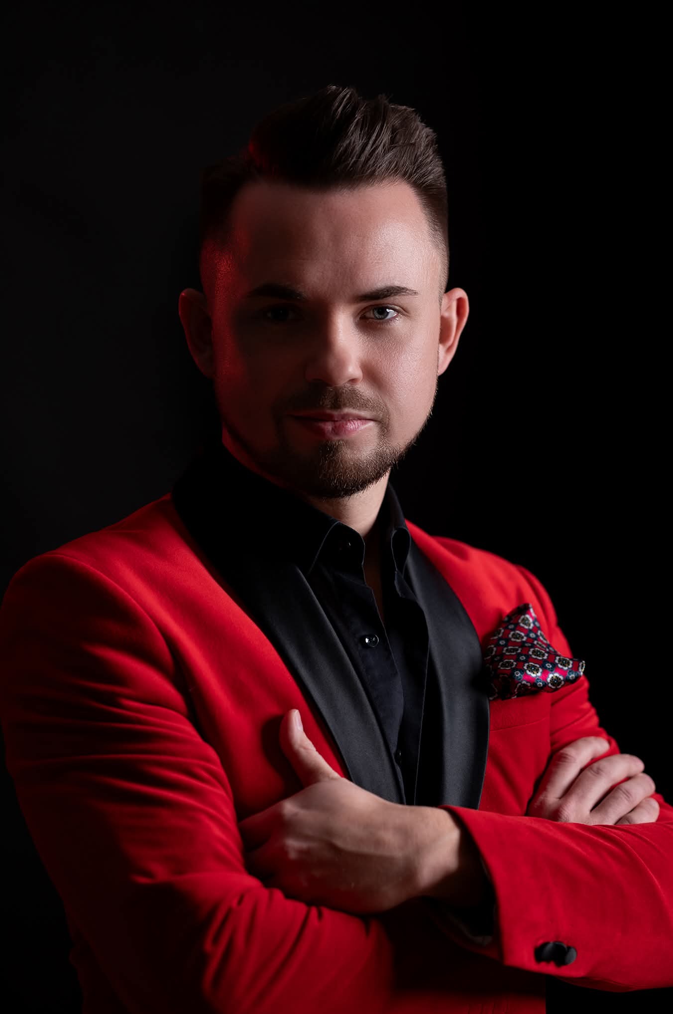 Portret mężczyzny w czerwonej marynarce i czarnej koszuli na czarnym tle, z rękami skrzyżowanymi na piersiach. Ujęcie studyjne, oświetlenie podkreśla detale twarzy.