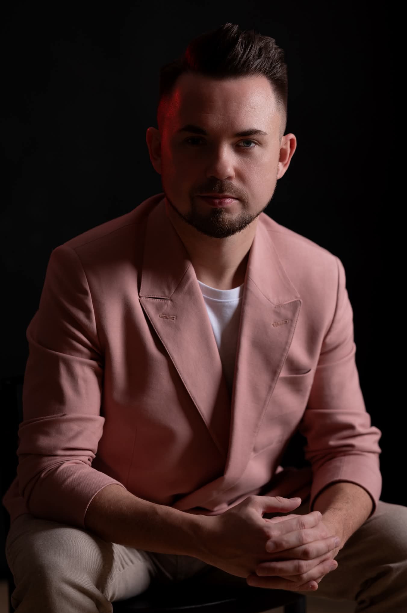 Portret mężczyzny w różowym garniturze na czarnym tle, siedzącego z dłońmi splecionymi. Czerwone oświetlenie na górze głowy, ciemne oczy, krótka broda.