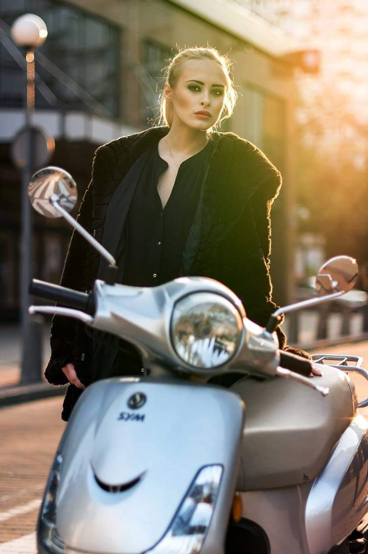 Blond modelka w czarnej kurtce pozuje na srebrnym skuterze SYM na tle miejskiej ulicy w ciepłym świetle zachodzącego słońca.