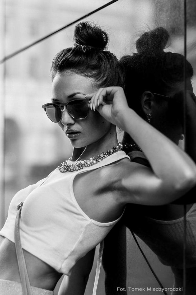 Czarno-białe ujęcie kobiety w okularach, opierającej się o szybę z odbiciem, z nonszalancko uniesioną ręką, prezentujące styl i elegancję.