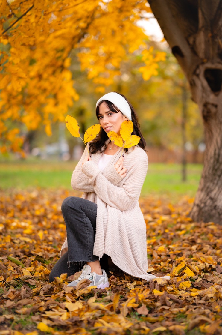 Jesienna sesja portretowa: brunetka w beżowym kardiganie i opasce, klęczy na tle drzewa, trzymając żółte liście. Ciepłe światło podkreśla złote barwy.