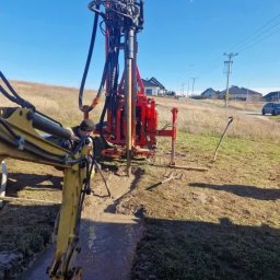 ZAINSTAL Usługi wodno-kanalizacyjne, wykończeniowe Małgorzata Pręgowska - Czerwona wiertnica w trakcie pracy, wspierana przez żółtą koparkę. W tle domy i błękitne niebo. Woda w wykopie. Prace ziemne w toku.