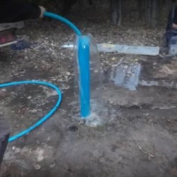 ZAINSTAL Usługi wodno-kanalizacyjne, wykończeniowe Małgorzata Pręgowska - Wypływająca woda z niebieskiej rury studni głębinowej, otoczonej ziemią i liśćmi, z widocznym wężem doprowadzającym wodę i fragmentem pojazdu w tle.