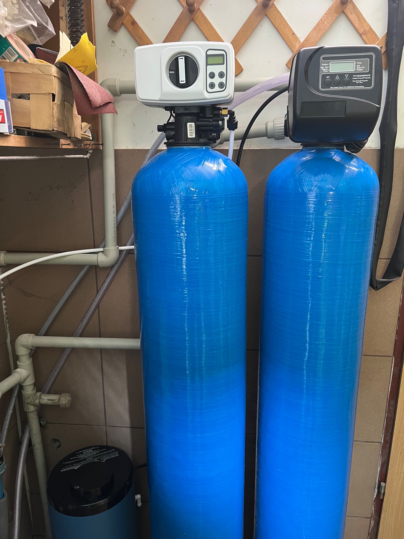 Dwa niebieskie filtry wody z elektronicznymi panelami sterowania, połączone rurami. Widoczne elementy instalacji wodnej i fragment ściany z drewnianym wieszakiem.