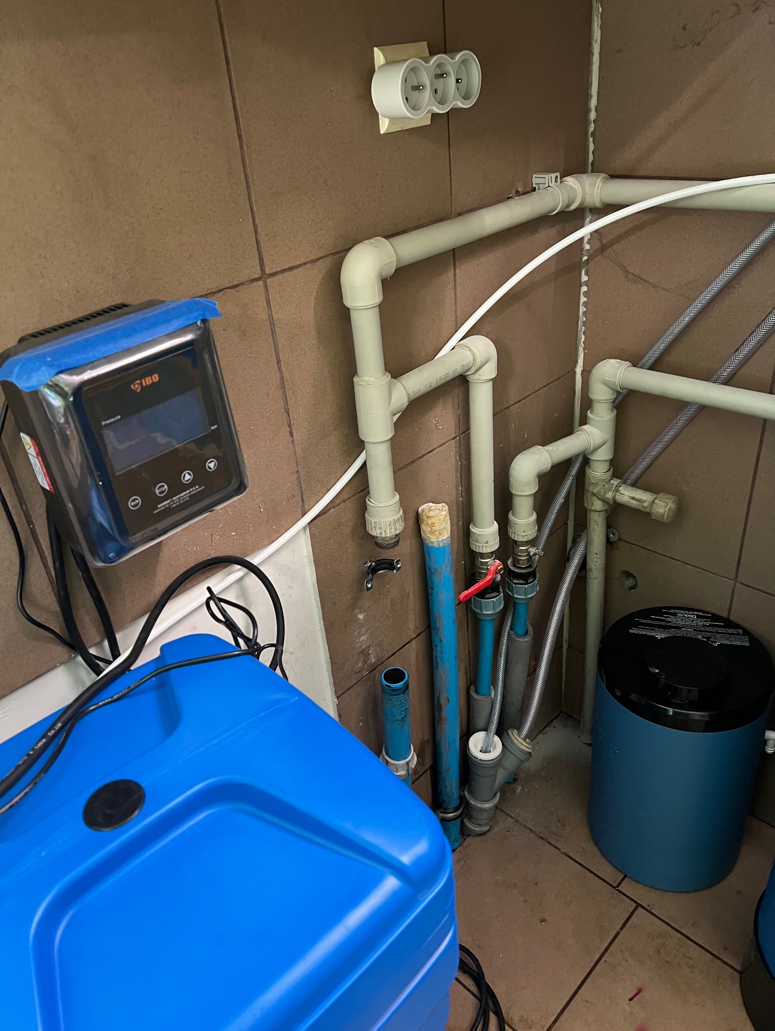 Zestaw hydroforowy IBO z niebieskim zbiornikiem i sterownikiem, widoczne rury instalacji wodnej na tle brązowych płytek. Elementy przyłączeniowe.