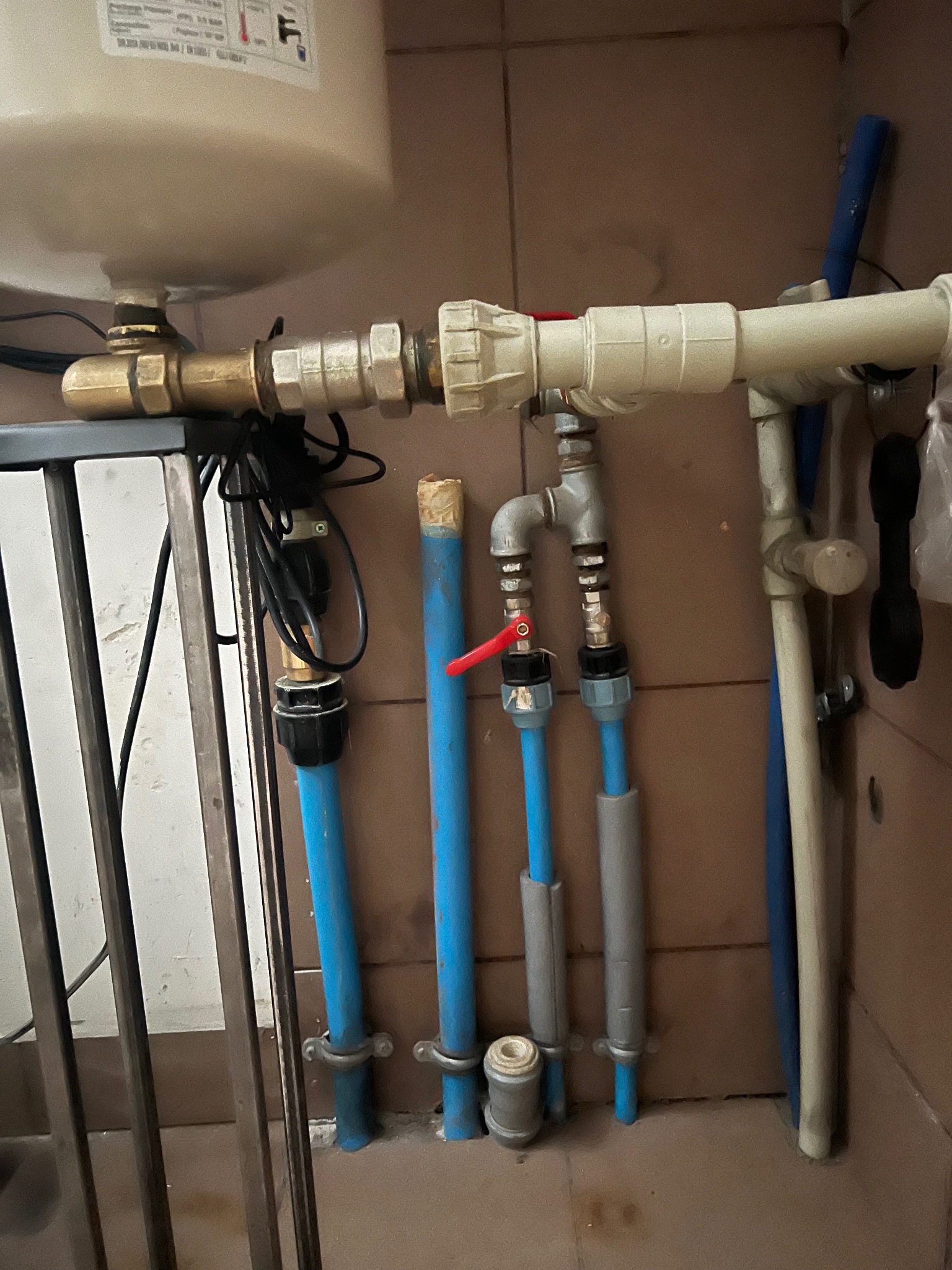 Kompleksowe przyłącze wodno-kanalizacyjne z rurami w kolorze niebieskim i szarym, z zaworem z czerwoną rączką, zamontowane przy ścianie z brązowych płytek.