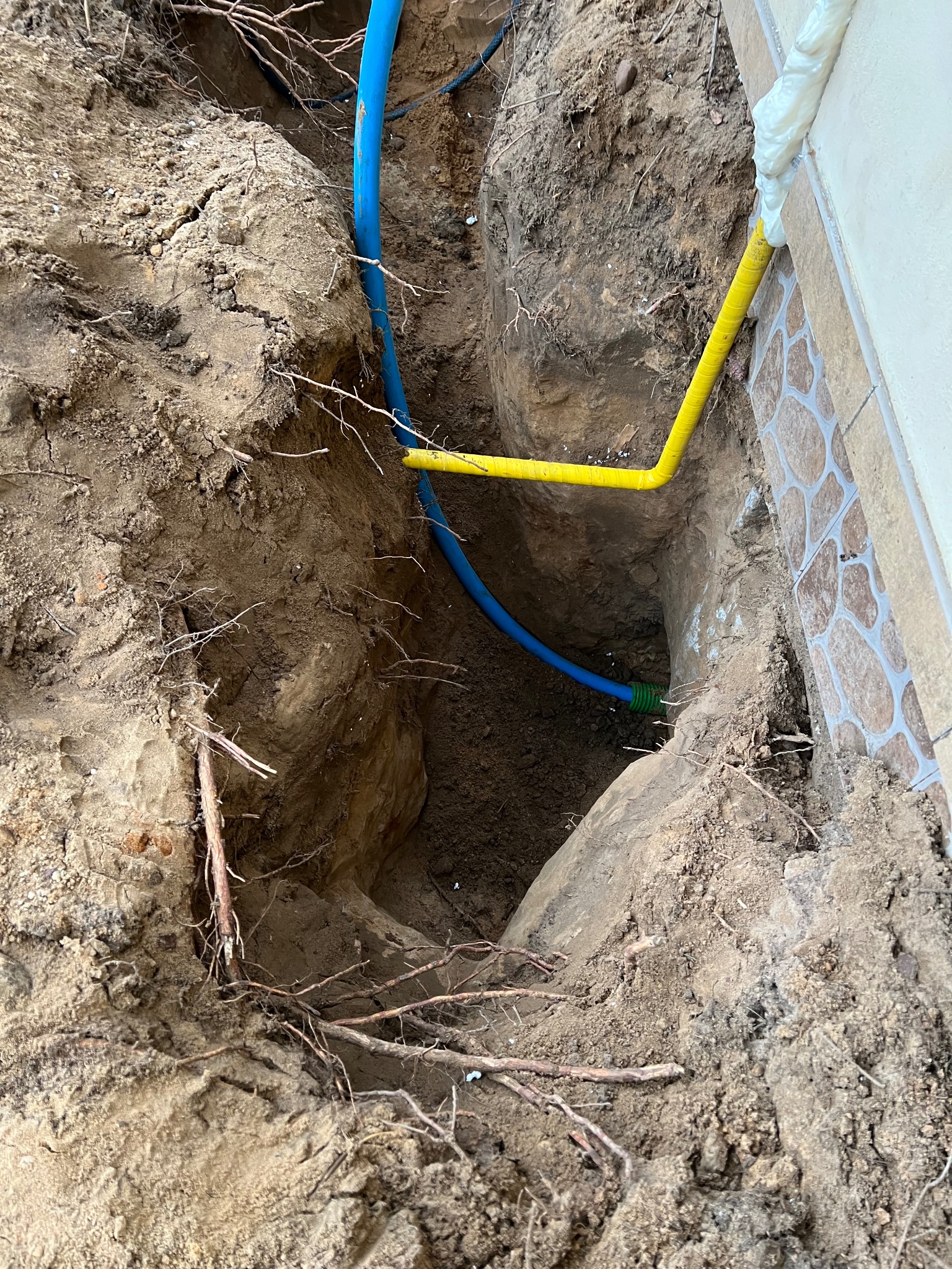 Wykop w ziemi z widocznymi rurami: niebieską i żółtą, prowadzącymi do budynku z dekoracyjną elewacją. Widoczne korzenie drzew. Prace instalacyjne w toku.