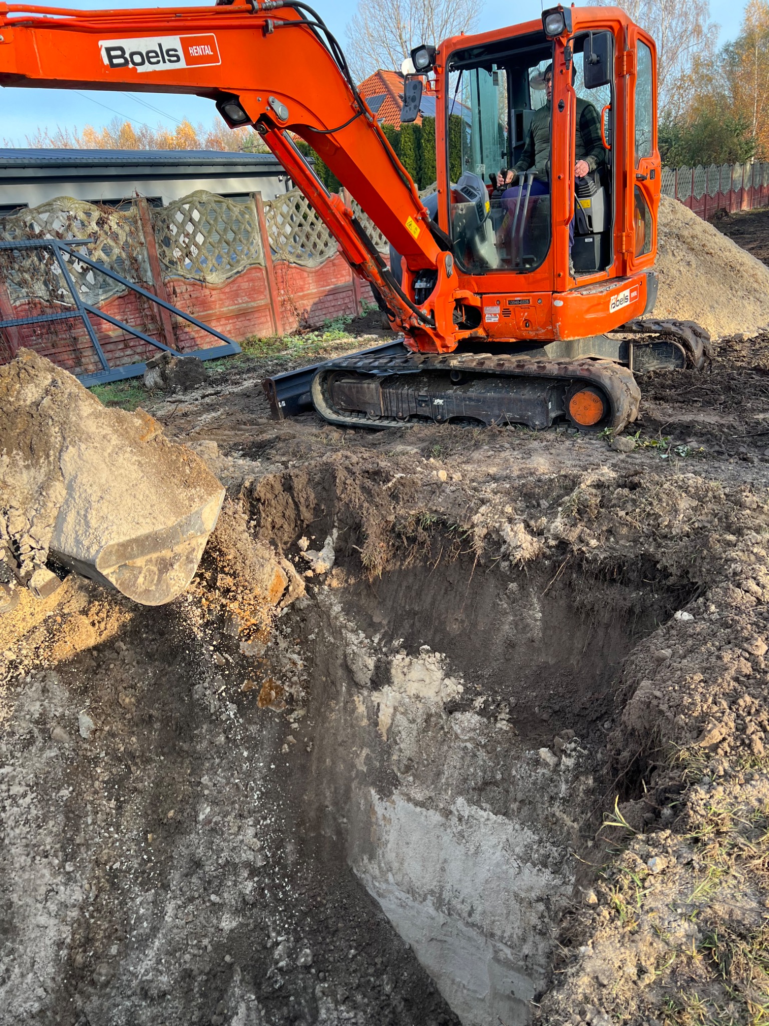 Pomarańczowa mini koparka Boels Rental w trakcie wykopu ziemi pod fundamenty. Widoczny operator w kabinie i wykopany dół. W tle ogrodzenie i drzewa.