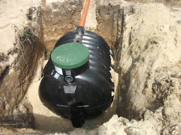 Czarny zbiornik z zieloną pokrywą w wykopie z piasku, prawdopodobnie przydomowa oczyszczalnia ścieków w trakcie montażu. Widoczne ściany wykopu.