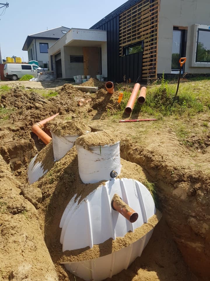 Biały zbiornik przydomowej oczyszczalni ścieków częściowo zasypany piaskiem w wykopie, z widocznymi rurami kanalizacyjnymi i budynkiem w tle.