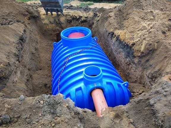 Niebieski zbiornik z rurą w wykopie, prawdopodobnie szambo betonowe lub przydomowa oczyszczalnia ścieków. Widoczny fragment koparki.