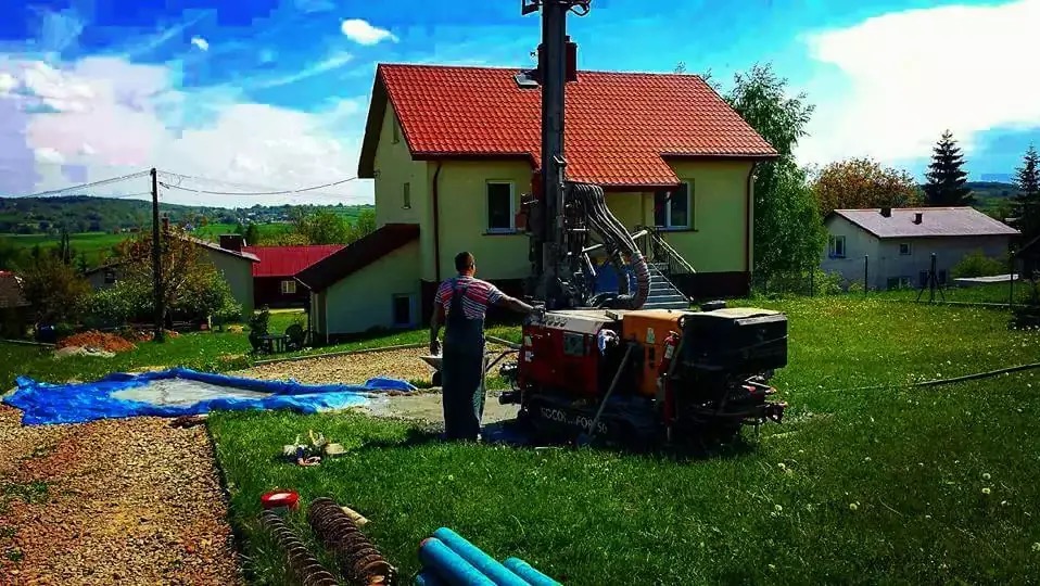 Prace wiertnicze na posesji: wiertnica w akcji na tle domu z czerwoną dachówką, pracownik nadzoruje proces, widoczne rury i sprzęt. Dzień słoneczny, zielone otoczenie.