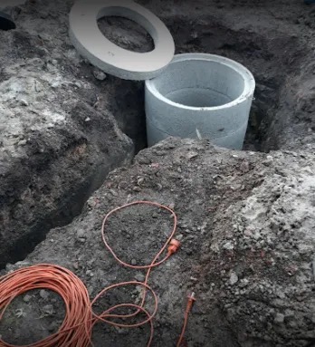 Betonowa studnia kręgowa w wykopie, z pokrywą obok. Pomarańczowy kabel na ziemi. Widok z góry na plac budowy.