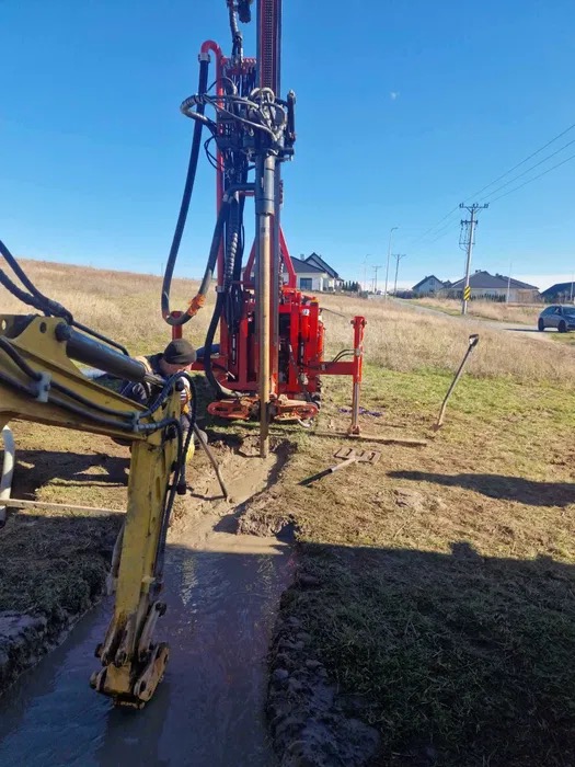 Czerwona wiertnica w trakcie pracy, wspierana przez żółtą koparkę. W tle domy i błękitne niebo. Woda w wykopie. Prace ziemne w toku.