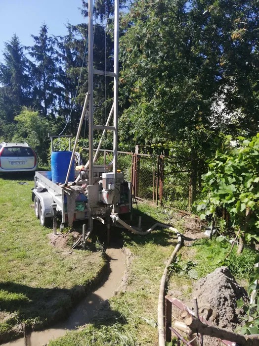 Wiertnica na przyczepie w trakcie pracy na trawiastym terenie, widoczny wykop i wąż odprowadzający wodę. W tle drzewa i samochód osobowy.