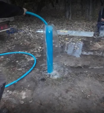 Wypływająca woda z niebieskiej rury studni głębinowej, otoczonej ziemią i liśćmi, z widocznym wężem doprowadzającym wodę i fragmentem pojazdu w tle.