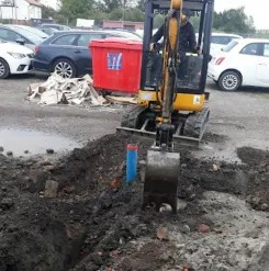 Żółta mini koparka w trakcie wykopu. W tle czerwony kontener, zaparkowane auta i budynki. Widoczny fragment niebieskiej rury w wykopie.