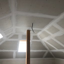 SMARTBUD - Poddasze z płytami gipsowo-kartonowymi, szpachlowane łączenia. Widoczne okna dachowe i drewniany słup podtrzymujący konstrukcję. Detal: czarny kabel zwisający z sufitu.