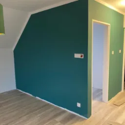 Pomieszczenie po malowaniu: ściana w odcieniach zieleni, białe wykończenia, drewnopodobna podłoga, widoczne gniazdka elektryczne i fragment zielonego abażuru lampy.