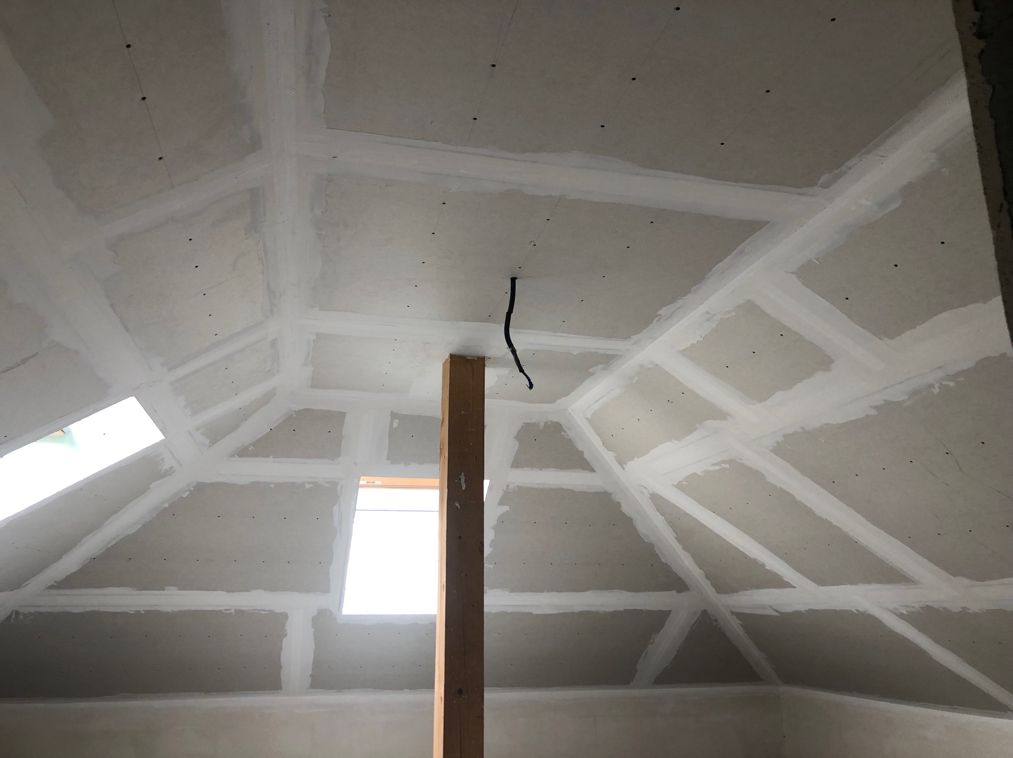 Poddasze z płytami gipsowo-kartonowymi, szpachlowane łączenia. Widoczne okna dachowe i drewniany słup podtrzymujący konstrukcję. Detal: czarny kabel zwisający z sufitu.
