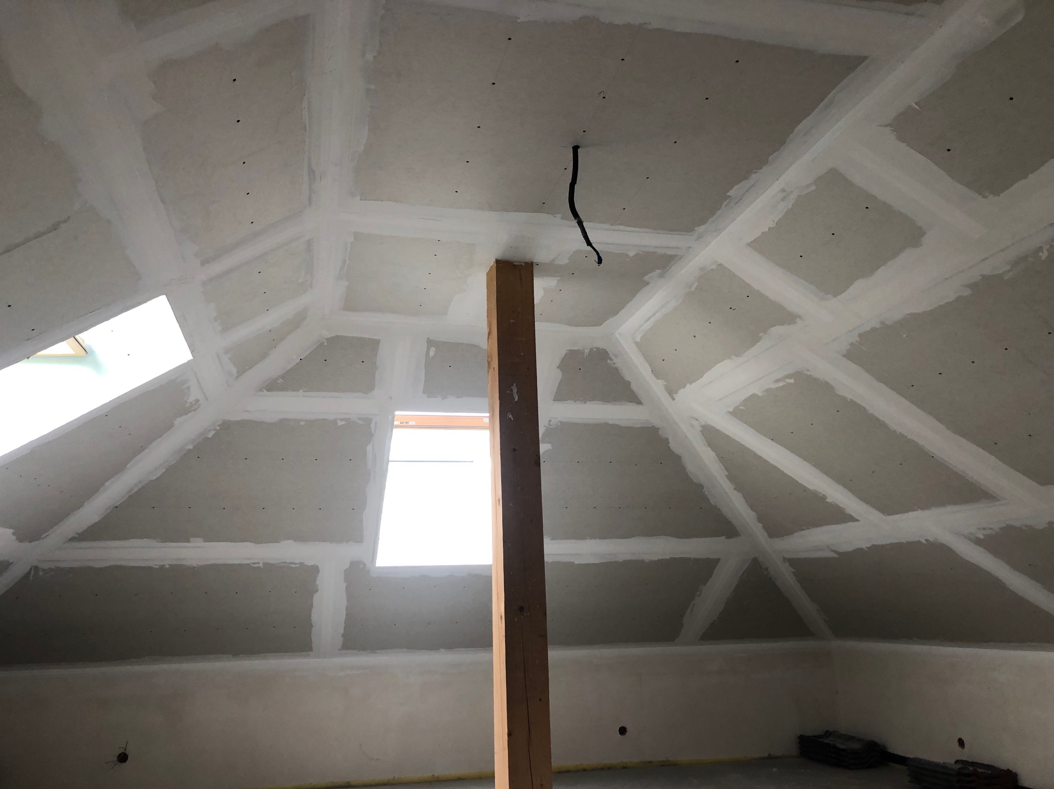 Poddasze po montażu płyt gipsowo-kartonowych z widocznymi spoinami, drewniany słup podtrzymujący konstrukcję, okno dachowe w skośnej ścianie, surowy stan.