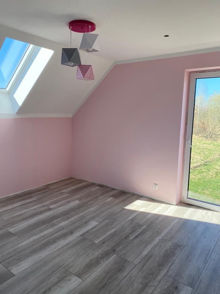 Pomalowane na różowo poddasze ze skosami i oknem dachowym, na podłodze panele imitujące drewno. Widoczny fragment lampy z geometrycznymi abażurami.