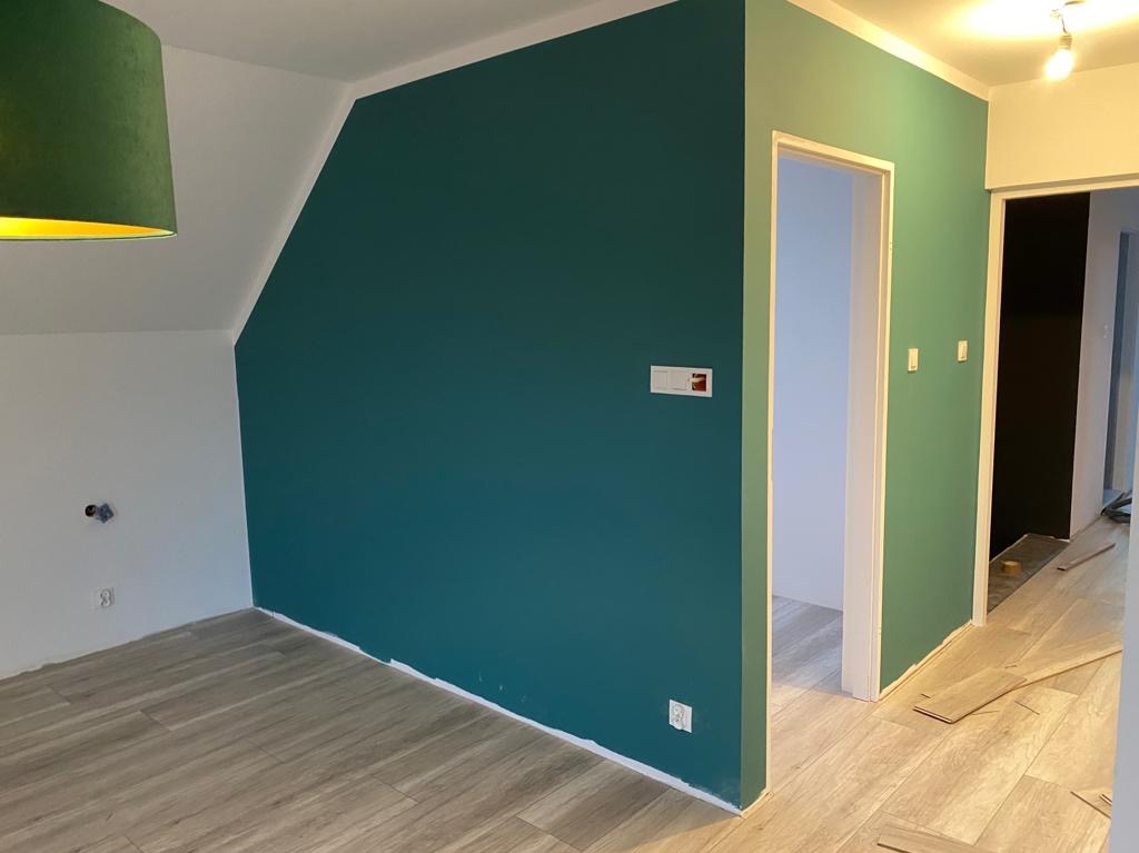 Pomieszczenie po malowaniu: ściana w odcieniach zieleni, białe wykończenia, drewnopodobna podłoga, widoczne gniazdka elektryczne i fragment zielonego abażuru lampy.