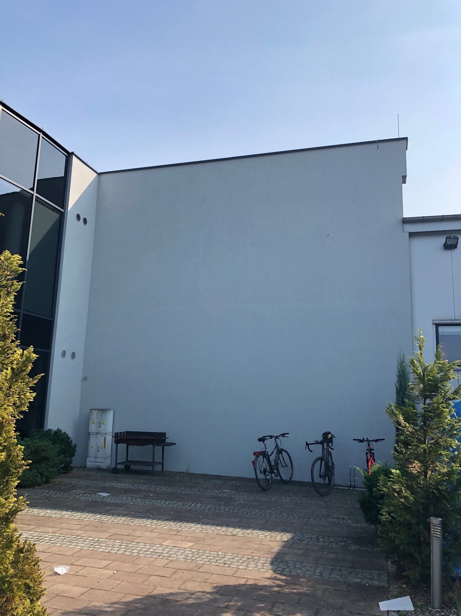 Jasnoszara fasada budynku z widocznymi śladami po szpachlowaniu, z trzema rowerami opartymi o ścianę i drzewami po bokach. Widoczne niedoskonałości w wykończeniu ściany.