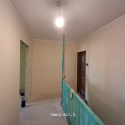 Mateusz Stopka - Jasne, świeżo pomalowane ściany w korytarzu z widocznymi listwami przypodłogowymi i zabezpieczeniem z folii malarskiej. Widoczne drzwi i fragment niezakończonego przejścia.