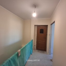 Mateusz Stopka - Jasne, świeżo pomalowane ściany korytarza z widoczną drewnianą ościeżnicą i drzwiami. Podłoga zabezpieczona folią malarską. Widoczne ślady prac wykończeniowych.