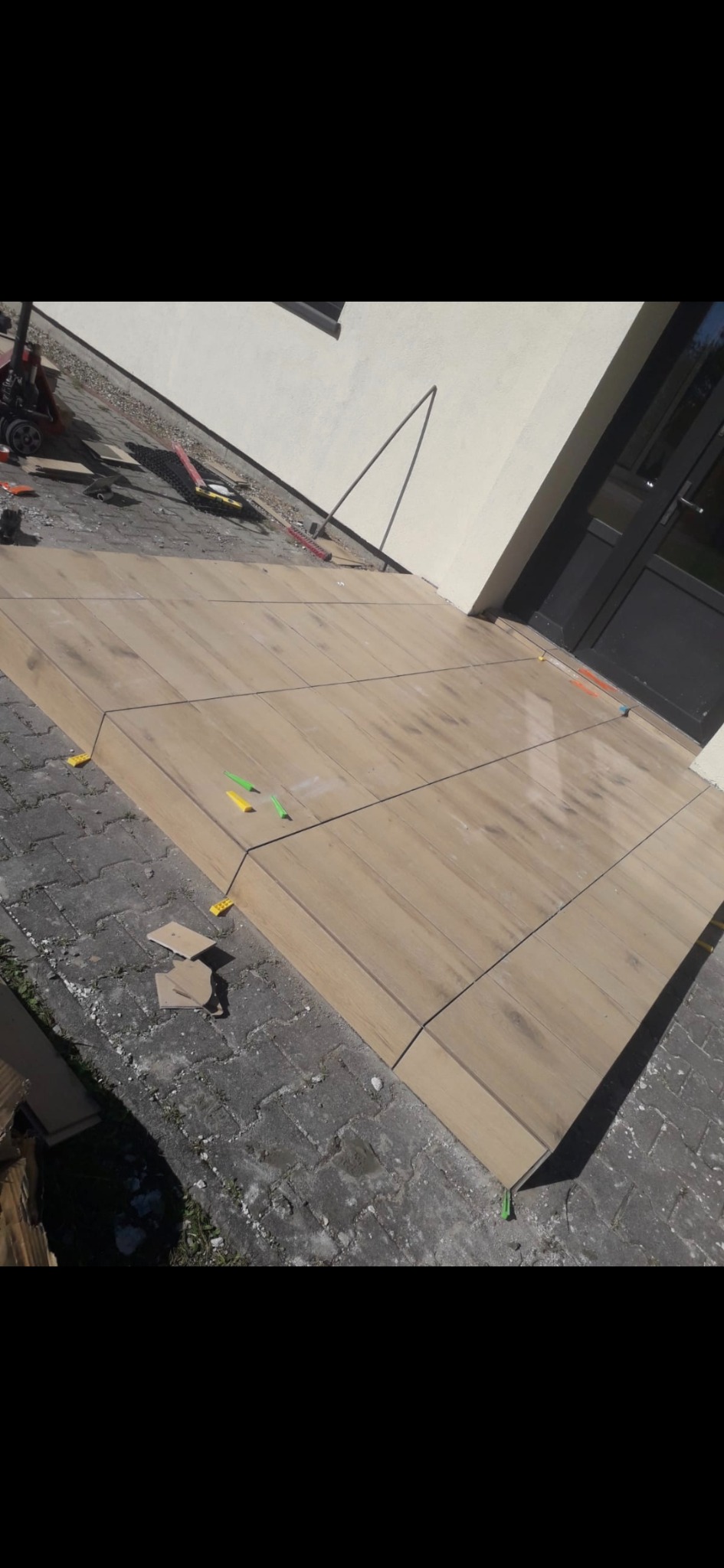 Układanie płytek imitujących drewno na tarasie, widoczne kliny poziomujące, narzędzia glazurnicze w tle. Etap prac wykończeniowych na zewnątrz budynku.