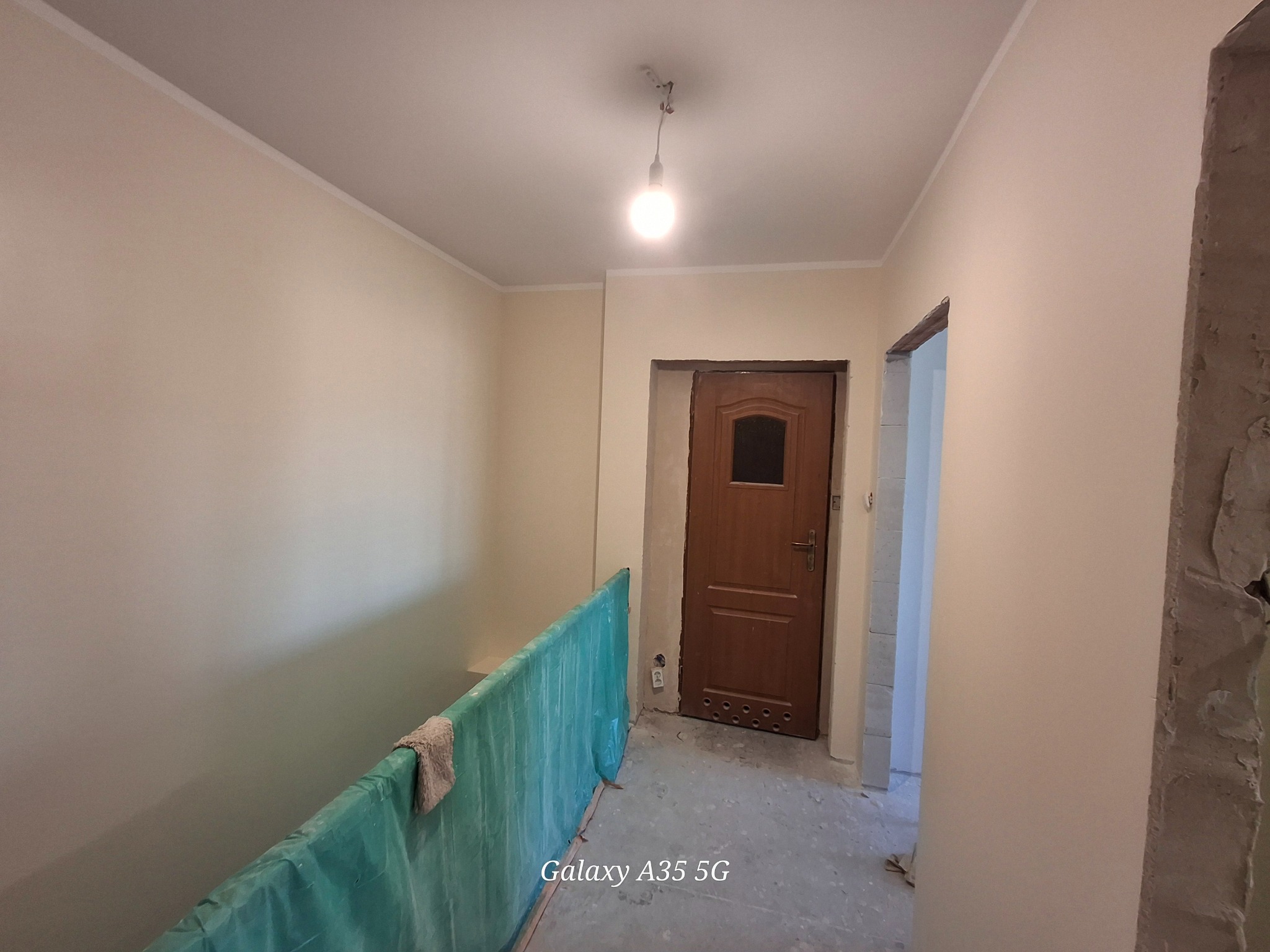 Jasne, świeżo pomalowane ściany korytarza z widoczną drewnianą ościeżnicą i drzwiami. Podłoga zabezpieczona folią malarską. Widoczne ślady prac wykończeniowych.