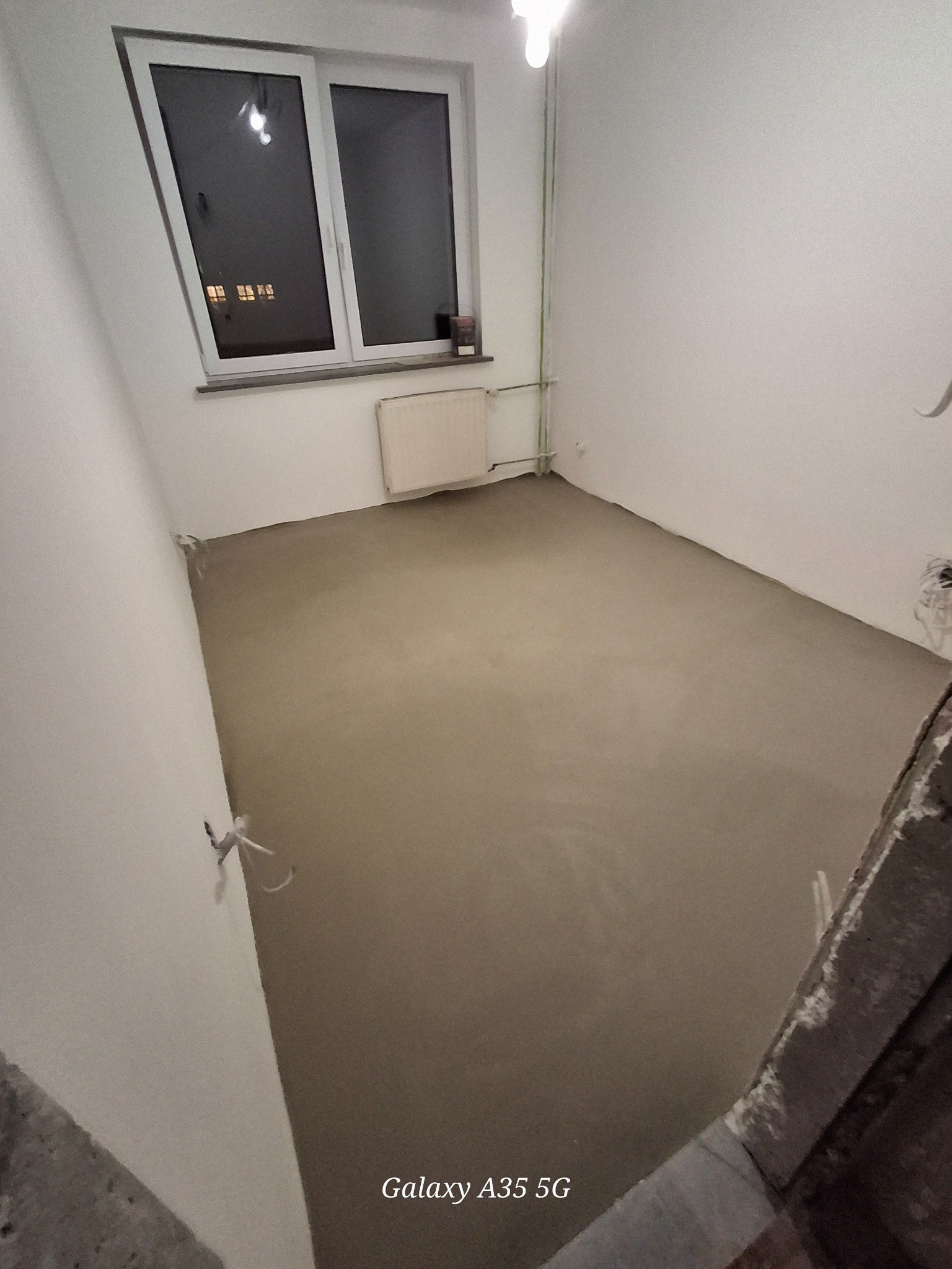 Wnętrze pokoju po wylaniu wylewki samopoziomującej. Widoczne gładkie podłoże, okno z parapetem i grzejnik. Ściany białe, przygotowane do dalszych prac wykończeniowych.