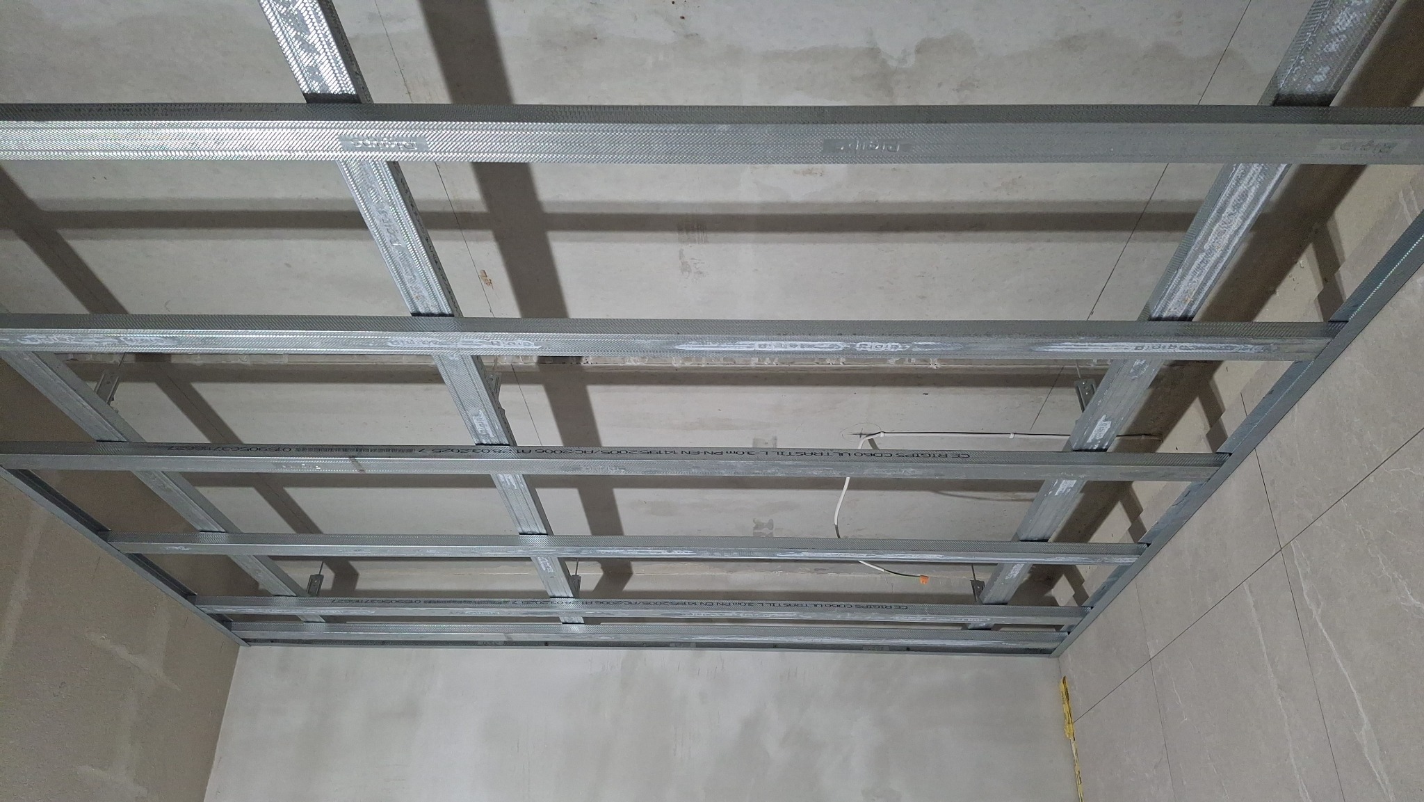 Metalowa konstrukcja sufitu podwieszanego z widocznymi profilami CD i UD, zamocowana do surowego stropu. Widoczne przewody elektryczne. Kąt ujęcia z dołu.
