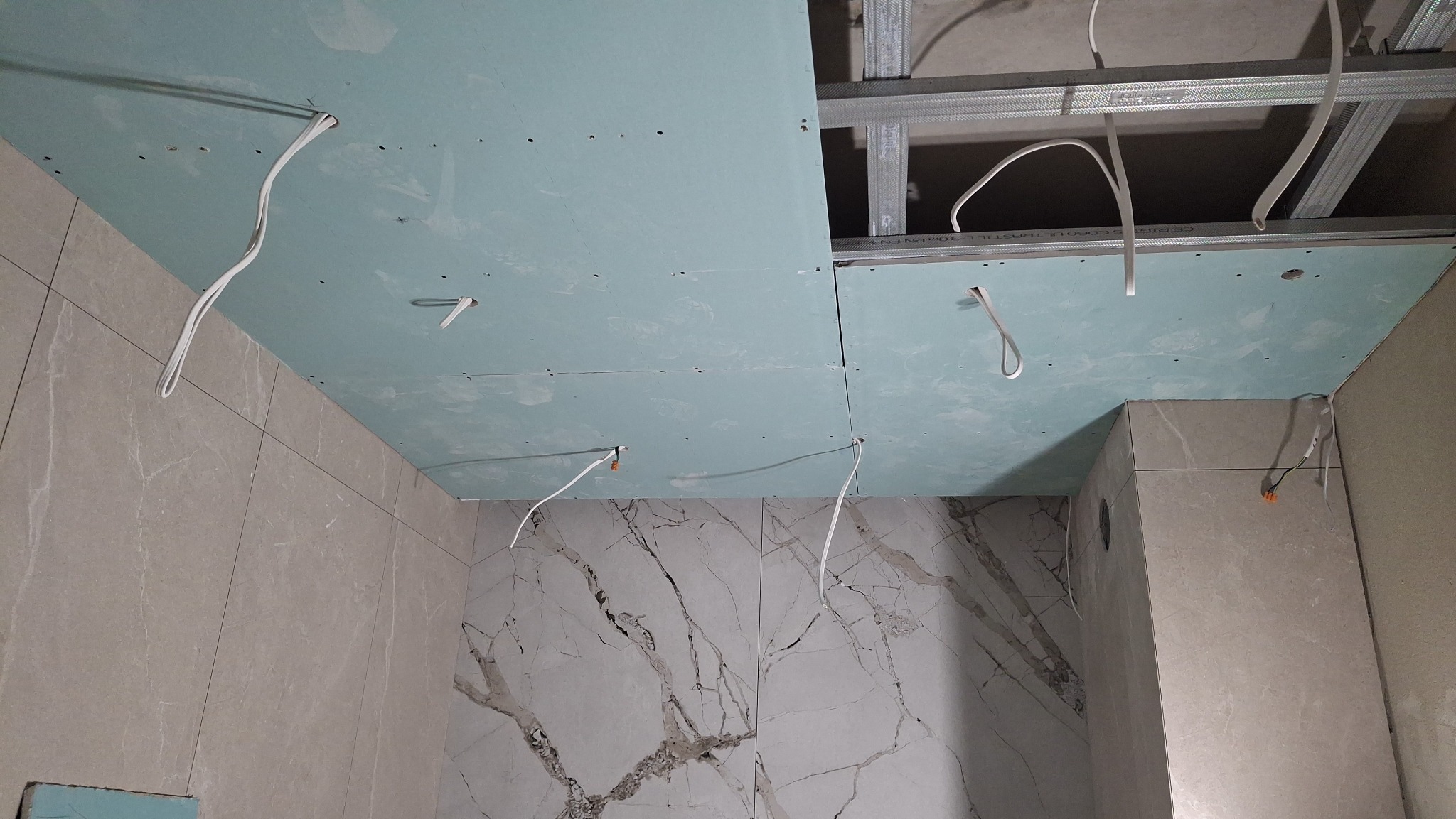 Niebieska płyta gipsowa na suficie z widoczną metalową konstrukcją i wystającymi kablami w łazience z marmurowymi płytkami na ścianach. Ujęcie z dołu.