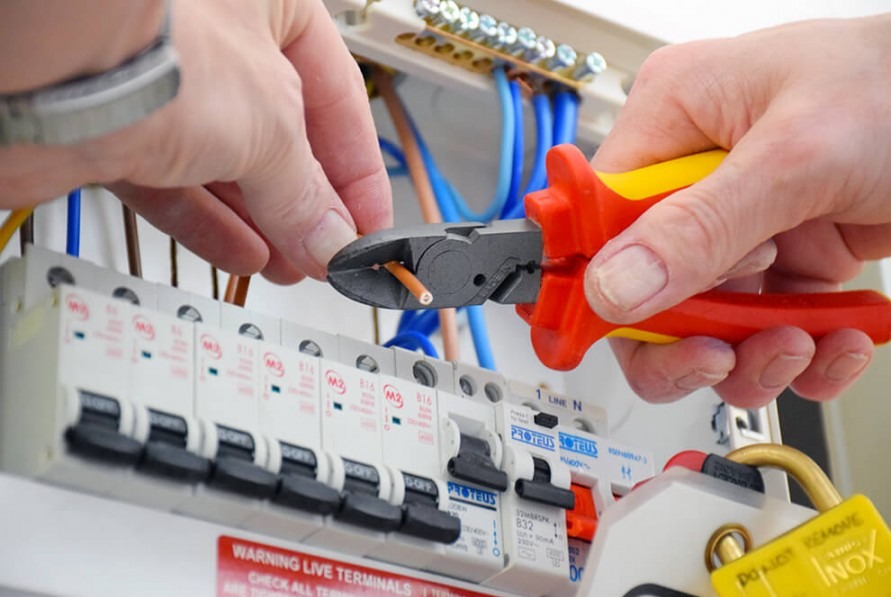 Elektryk obcina przewód w skrzynce bezpiecznikowej kombinerkami z żółto-czerwoną rękojeścią. Widoczne zabezpieczenia i kable w różnych kolorach.