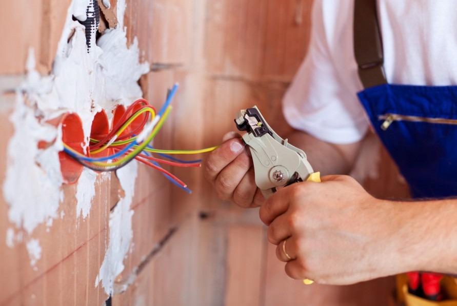 Elektryk w trakcie pracy: przygotowanie przewodów w puszce instalacyjnej w ścianie z cegły, używając ściągacza izolacji. Widoczne przewody w kolorach: niebieski, żółty, czerwony.