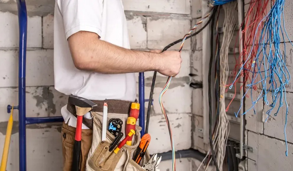 Elektryk w trakcie pracy z kablami w surowym wnętrzu. Widoczny pas z narzędziami, drabina i ściana z betonu komórkowego.